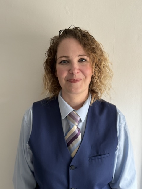 Lucy with light brown curly hair, wearing a pale blue shirt, navy waistcoat and blue striped tie, smiling against a cream background.