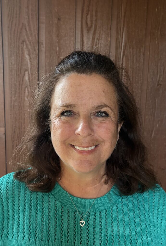 A woman with brown hair, smiling and wearing a turquoise shirt, standing in front of a brown wooden wall.
