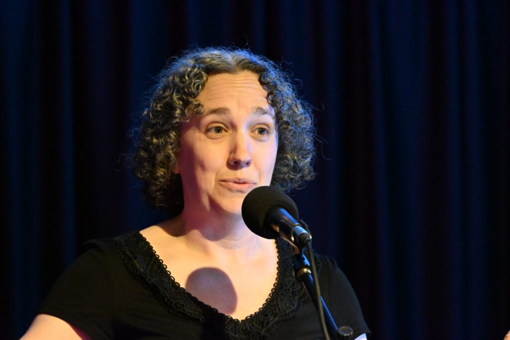 A photograph of Sam singing into a microphone on a stage. She has brown curly hair with grey highlights and is wearing a black top with lace edging.