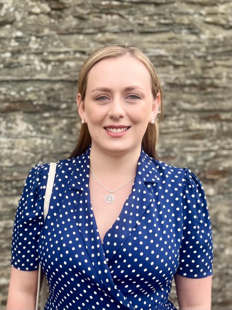 Professional headshot of a woman wearing a blue polka-dot dress and silver necklace, smiling at the camera in front of a stone wall.